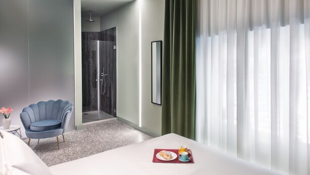 Ein modernes Hotelzimmer mit einem Bett, einem blauen Sessel, einem kleinen Tisch mit Teller und Tasse sowie einer Dusche.