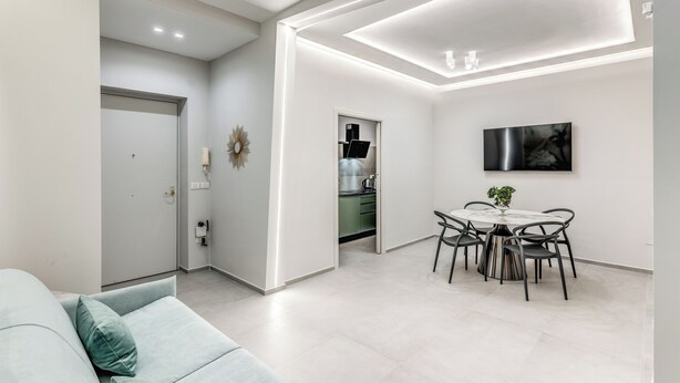 Una sala de estar moderna con un sofá, una mesa de comedor con sillas, una cocina al fondo e iluminación empotrada en el techo.