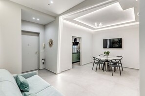 Ein modernes Wohnzimmer mit einer Couch, einem Esstisch mit Stühlen, einer Küche im Hintergrund und Einbaustrahler an der Decke.