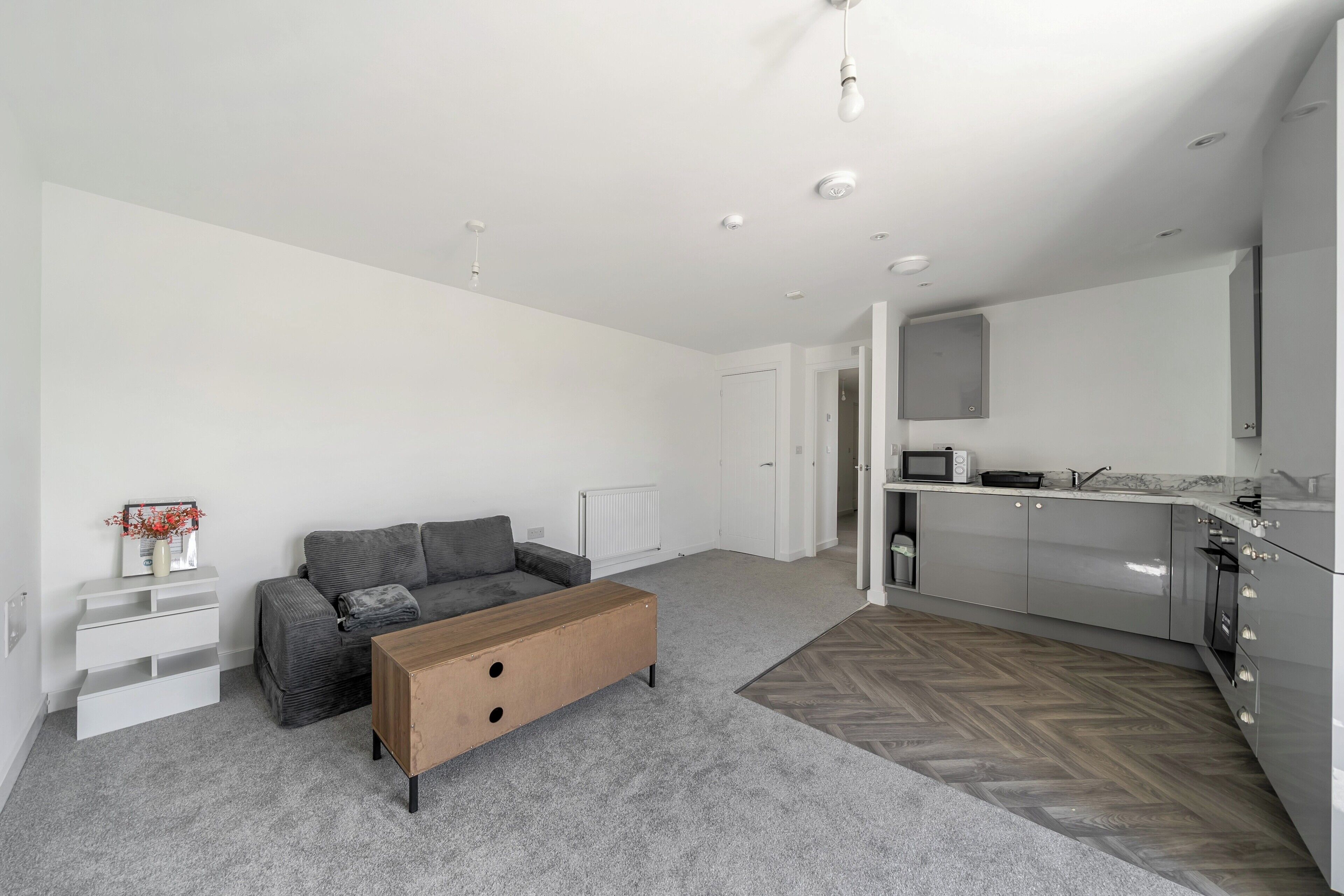 Een moderne woonkamer met een grijze bank, een houten salontafel en een keuken met grijze kastjes en een magnetron.