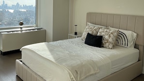 A modern bedroom with a large bed, a bedside table, a lamp, and a view of a cityscape through a window.