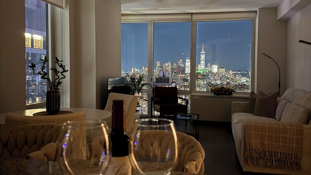A modern living room with a city view, a glass-top table with wine glasses and a bottle, and comfortable seating.