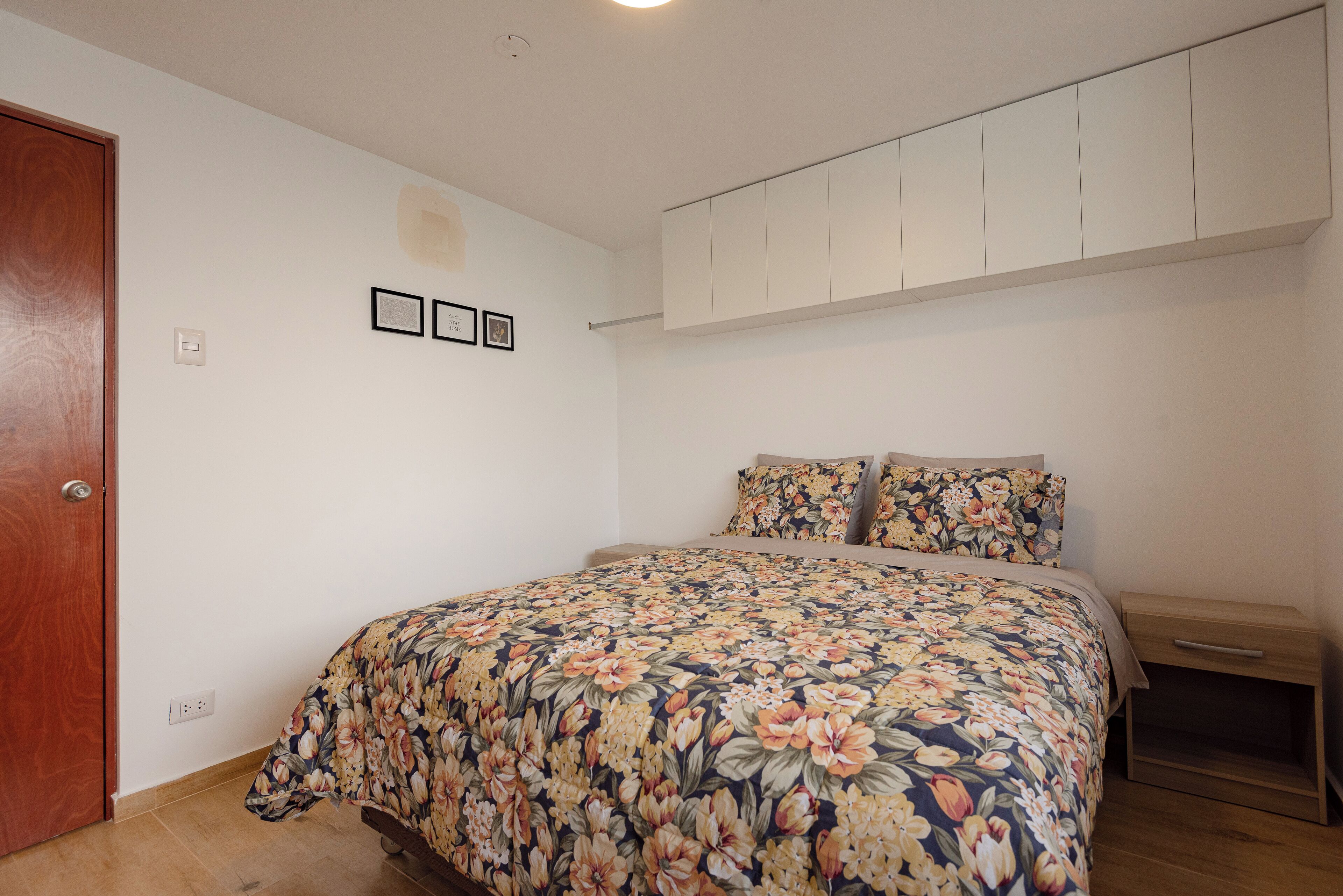 Une chambre à coucher avec une couvre-lit à motifs floraux, une porte en bois, un mur blanc orné de tableaux encadrés et un placard intégré au-dessus du lit.