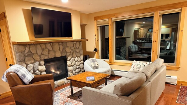A cozy living room with a stone fireplace, a flat-screen TV mounted above it, and a large window offering a view of the kitchen.