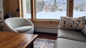A cozy room with a large window, a wooden table, two grey armchairs, and a decorative pillow.
