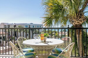 Ein runder Tisch mit Korbstühlen und Kissen, einer Flasche SUN POP sowie zwei Weingläsern auf einem Balkon mit Blick auf Gebäude und Palmen.