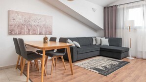 Uma sala de estar moderna com área de jantar, apresentando uma mesa e cadeiras de madeira, um sofá cinza com almofadas, um lustre pendente e um tapete com tema urbano.