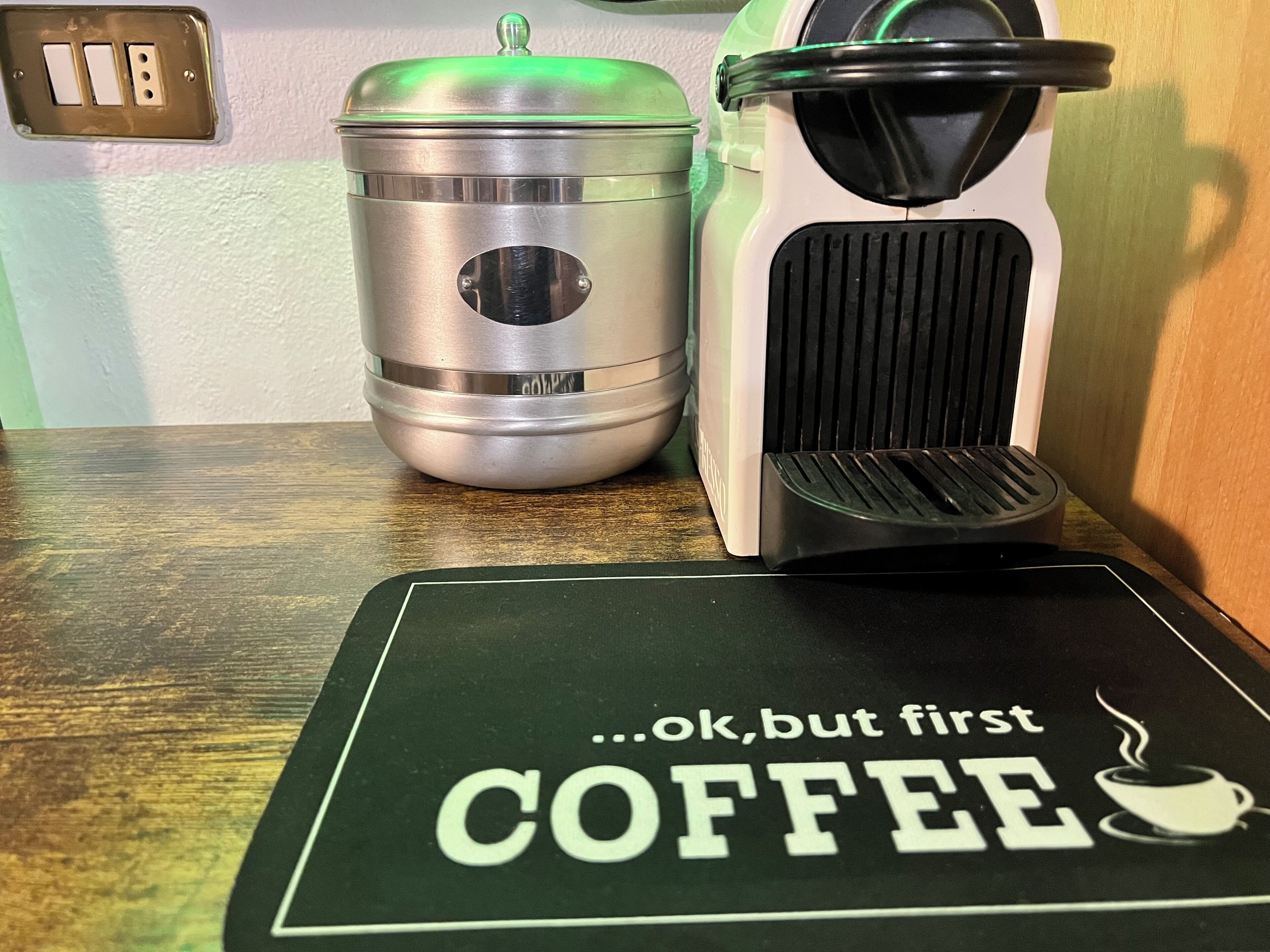 Un piano di lavoro della cucina con una macchina da caffè, un contenitore in acciaio inossidabile e un sottopiattino verde con la scritta ‘…ok, ma prima CAFFÈ’.