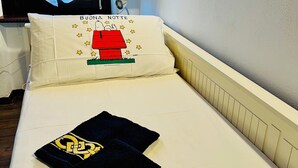 Habitación de hotel con cama, almohada con un personaje de dibujos animados, mesita de noche con una botella y ventana con cortinas.