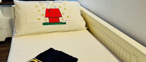Een hotelkamer met een bed, een kussen met een stripfiguur, een nachtkastje met een fles en een raam met gordijnen.
