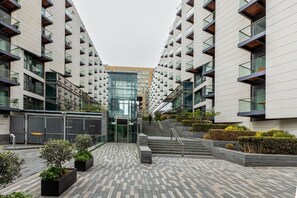 Ein modernes Gebäude mit einem zentralen Glaszugang, umgeben von einem gepflasterten Innenhof mit Pflanzgefäßen und Stufen zum Eingang.