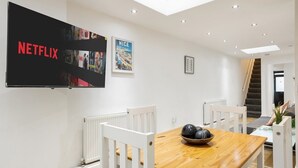 Een moderne eetruimte met een houten tafel, witte stoelen, een fruitschaal en een grote flatscreen-tv aan de muur met het Netflix logo.