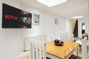 A modern dining area with a wooden table, white chairs, a bowl of fruit, and a large flat-screen TV mounted on the wall displaying the Netflix logo. - Modern Spacious Greenwich 2BR Central Location (Greater London)