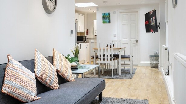 Een moderne woonkamer met een grijze bank, gekleurde kussens, een eethoek met een witte tafel en stoelen, een keuken op de achtergrond en een wandgemonteerde televisie.