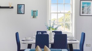 Uma área de refeições com mesa branca, cadeiras estofadas em azul e uma janela grande com vista para o verde.