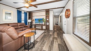 A living room with a brown leather sofa, a wooden coffee table, a ceiling fan, and a view of the outdoors through large windows. - Joy Drive In Movie & RV Resort - 2995 Barr Ln - Relaxing 3BR/2BA Getaway (Copperas Cove)