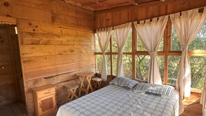 Ein Schlafzimmer einer Holzhütte mit einem Bett, einem kleinen Holztisch und einem Fenster mit Vorhängen.