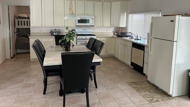 Une cuisine compacte avec des meubles blancs, un four à micro-ondes, une cuisinière et un réfrigérateur. On y trouve une table à manger avec des chaises et une plante en pot.