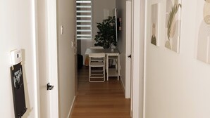 A narrow hallway with wooden flooring, white walls, and a ceiling light. There are doors on both sides, a small table with chairs, and a potted plant in the background. - suite, Cozy & Fully Equipped. (Edmonton)