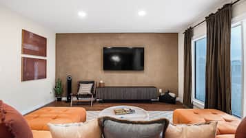 A living room with a large flat-screen TV mounted on the wall, a sofa set, a coffee table, and a window with curtains.