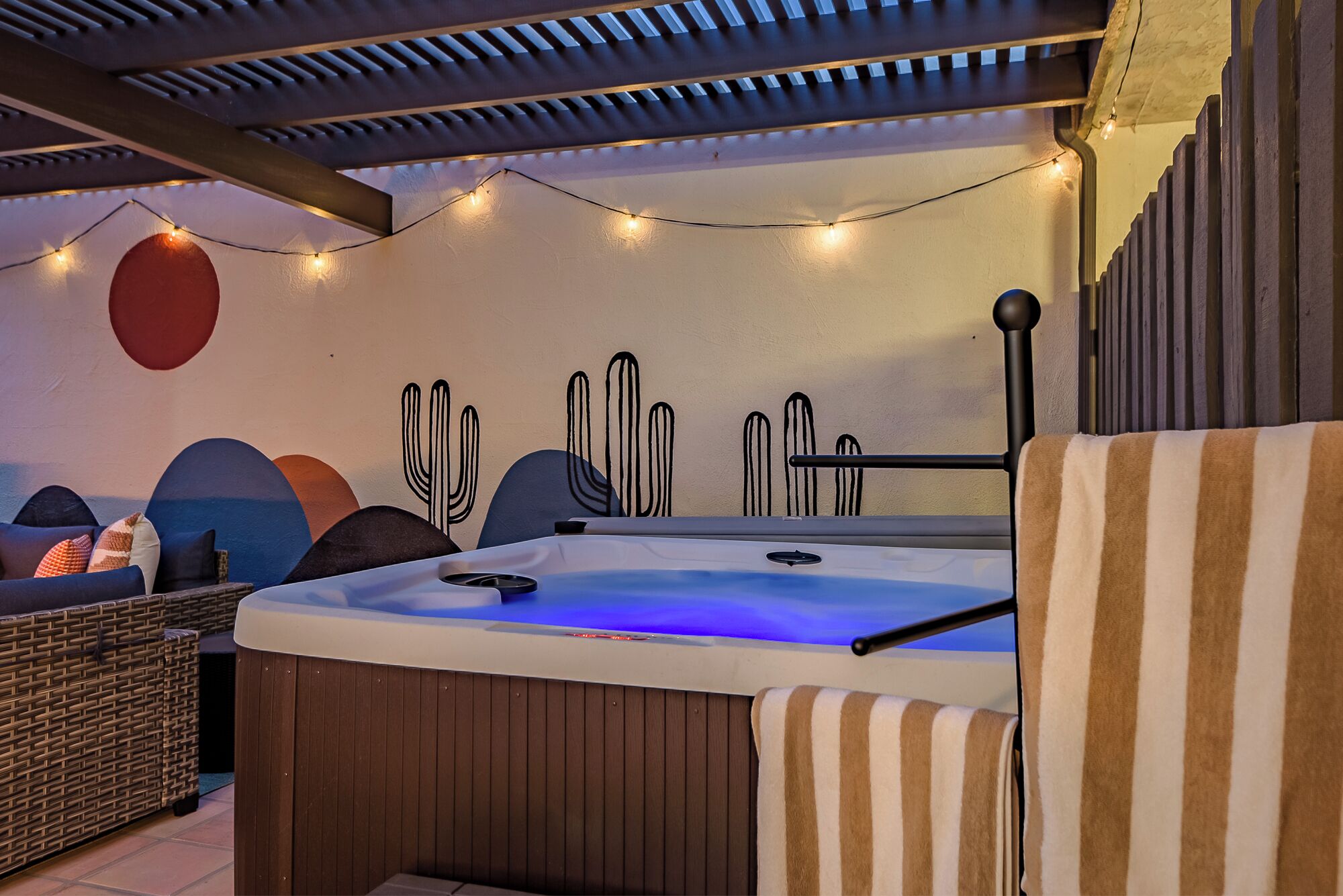 A hot tub with a wooden base and a blue light, surrounded by wicker furniture and a wall with cacti designs.