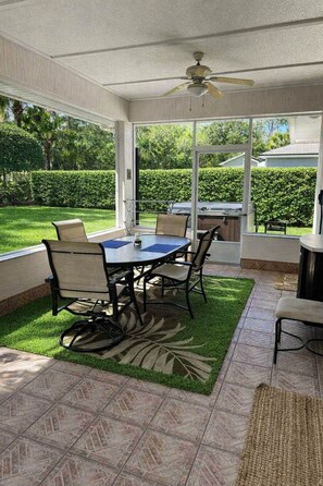 Un patio cubierto con una mesa y sillas, un ventilador de techo y vistas a un jardín cuidado.