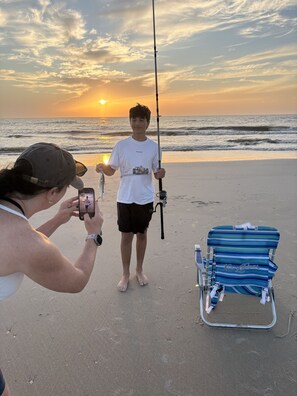 A person standing on a beach holding a fishing rod with a sunset in the background.