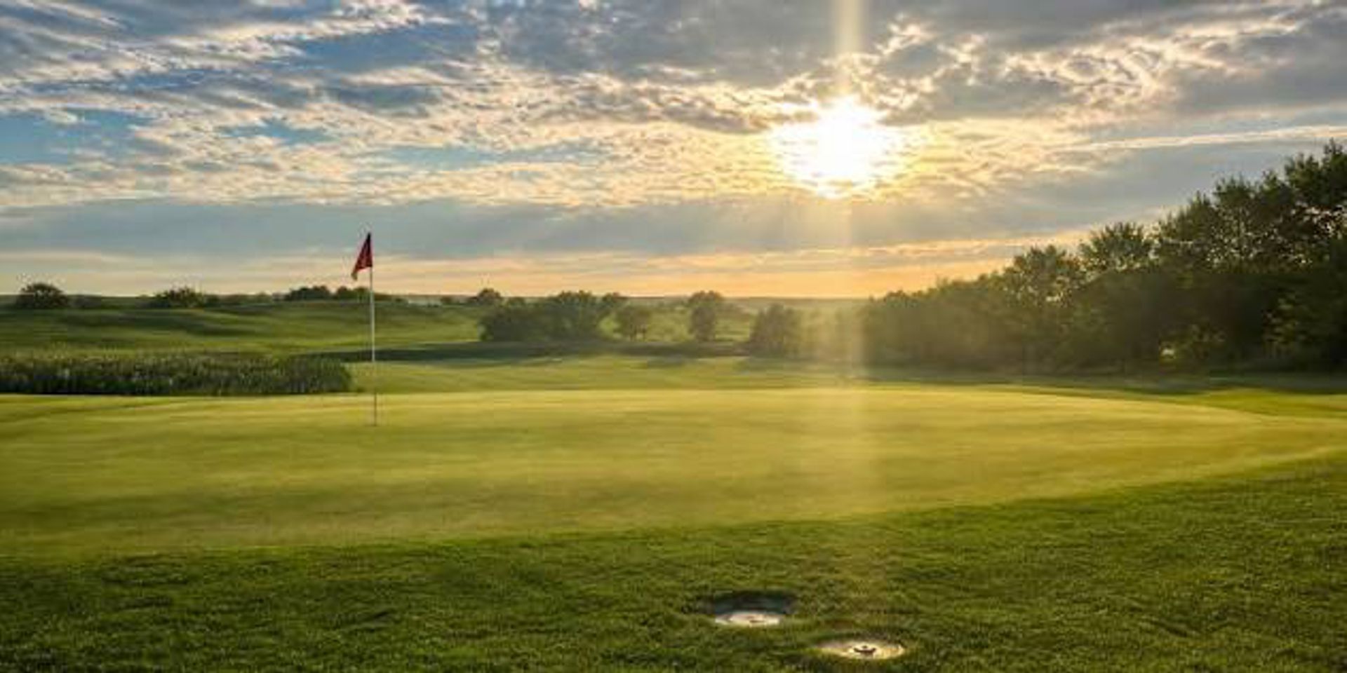 Un terrain de golf avec un drapeau, des arbres et un coucher de soleil.