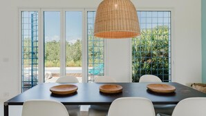 A dining area with a table, chairs, a hanging light fixture, and large windows with a view of greenery. - Villa Zabbara, independent and private pool (CASTELLAMMARE DEL GOLFO)