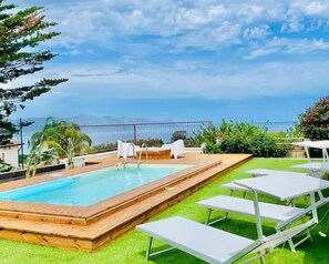 Piscina retangular com deck de madeira, rodeada por espreguiçadeiras brancas e um gramado.