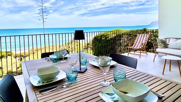 Mesa de jantar posta com copos e pratos, com vista para uma praia e céu claro.
