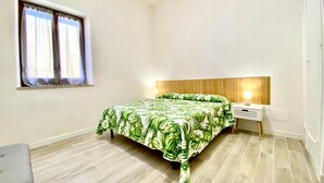 Quarto com cama, mesa de cabeceira, abajur, janela com cortinas e uma cadeira.