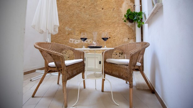 Eine Sitzecke mit Winkelsitz, Korbstühlen und einem kleinen Tisch, zwei Gläsern Rotwein und einem weißen Sonnenschirm.
