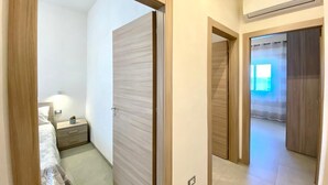 A bedroom with a bed, bedside table, and a nightstand with a lamp. There are wooden doors and a hallway leading to another room. - Villa dei Gerani, 500 mt from the sea. (ALCAMO)