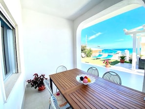 Ein Balkon mit einem Holztisch, Stühlen und einer Schale mit Obst. Im Erdgeschoss steht ein Blumentopf und man sieht einen Pool sowie eine Außenlounge.