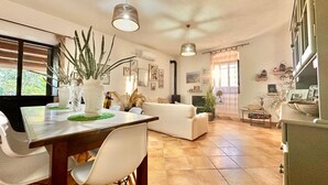 A spacious living area with a dining table, white chairs, a sofa, and a TV. There are plants, framed pictures, and a window with curtains. - Casale Bagni Villa with private and independent swimming pool (CANICATTINI BAGNI)