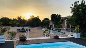 A swimming pool with a wooden deck, surrounded by a garden with various plants and a view of a desert landscape. - Casale Bagni Villa with private and independent swimming pool (CANICATTINI BAGNI)