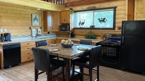 L’intérieur d’un chalet en bois comprend une cuisine équipée d’une table à manger, de chaises, d’un réfrigérateur et d’une fenêtre donnant sur la forêt.