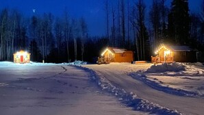 Schneebedeckte Landschaft mit drei Holzhütten, jede mit einer Veranda und warm beleuchtet.