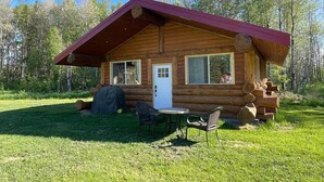 Punaisella katolla varustettu hirsihökkelö, jossa on etuovi ja pieni kuisti, jossa on kaksi tuolia ja pöytä.