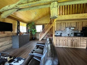 A rustic cabin interior with a leather sofa, wooden floors, a kitchen with wooden cabinets, and a staircase. - The Ram cabin - Bear Creek cabins (County Of Wetaskiwin No. 10)