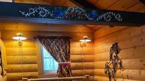 A log cabin bedroom with a bed, two wall-mounted lamps, a window with curtains, and a decorative wall piece. - The Mustang Cabin - Bear Creek Cabins (County Of Wetaskiwin No. 10)