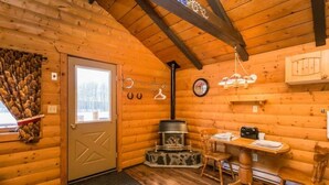 A cozy log cabin interior with a wooden door, a fireplace, a dining table with chairs, and a hanging light fixture. - The Mustang Cabin - Bear Creek Cabins (County Of Wetaskiwin No. 10)