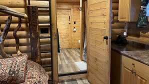 Interior de una cabaña de troncos con piso de madera, una cama con ropa de cama estampada, una puerta de madera que da a un baño y una pequeña zona de kitchenette.