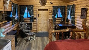 Interior de una cabaña de troncos con una mesa de comedor, un sillón estampado, una cama roja y techo de madera.
