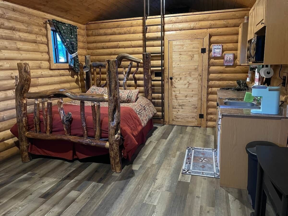 Quarto de cabana rústica com cama de madeira, um pequeno espaço para cozinha e janela com cortinas.