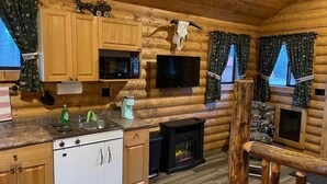 Interior de una cabaña de troncos con paredes de madera, una cocina con estufa y microondas, una chimenea y un televisor montado en la pared.