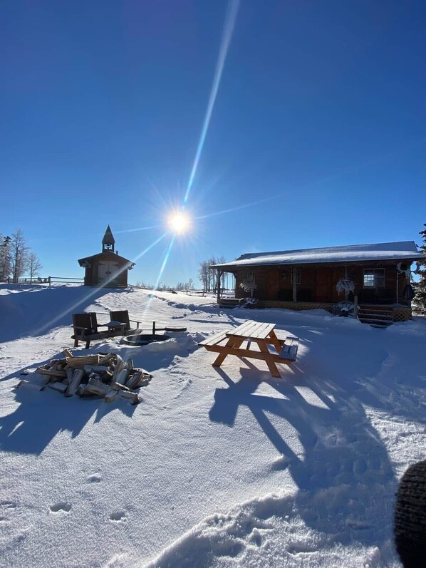 Una cabaña de madera con techo cubierto de nieve, una pequeña torre con campana y una mesa de picnic en primer plano.