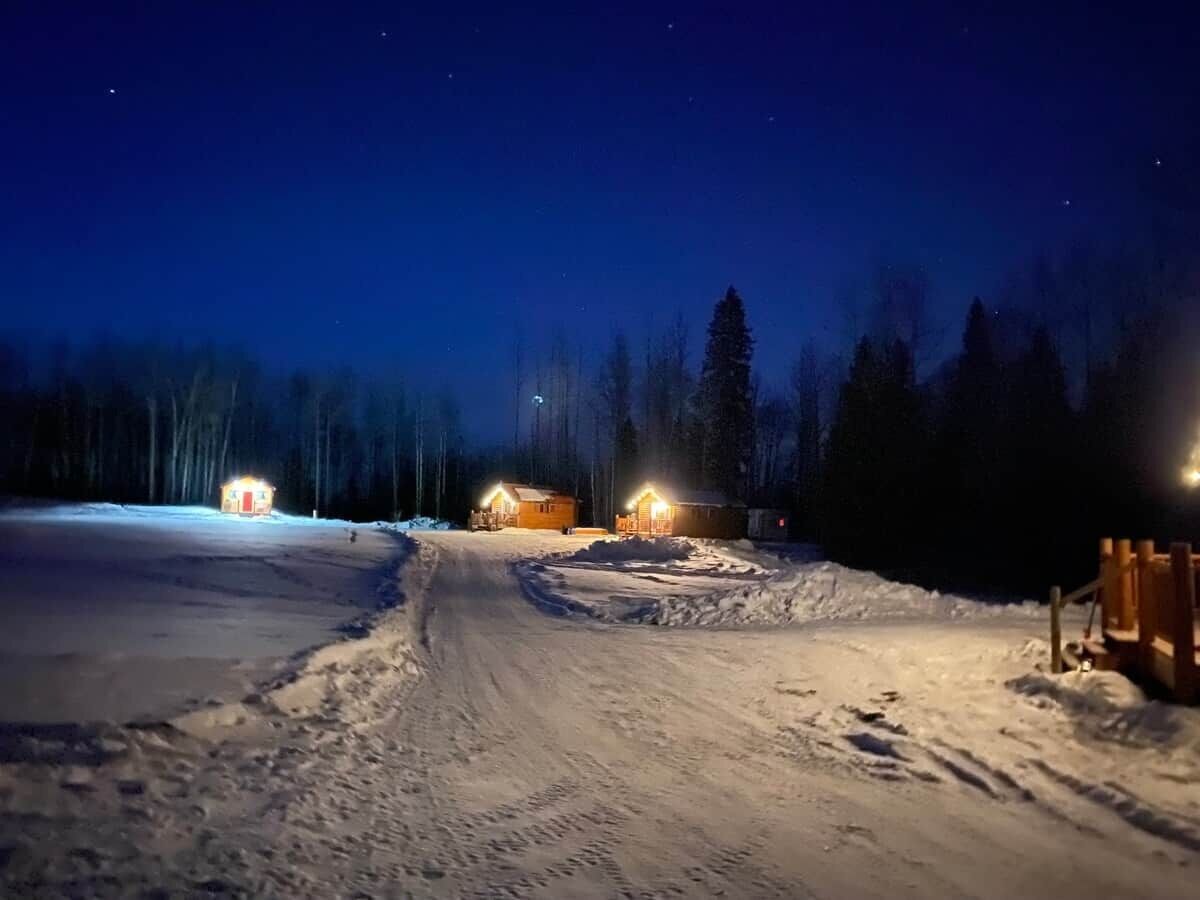 A snow-covered path leading to wooden cabins with lights, surrounded by trees.