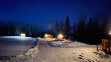 A snow-covered path leading to wooden cabins with lights, surrounded by trees.